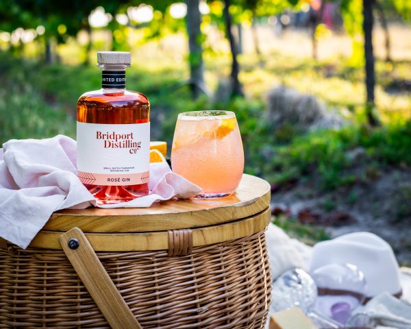 Bottle of Bridport Distilling Co's limited edition Rosé Gin sat in a picnic setting with a cocktail made from the gin with vineyard as the background setting